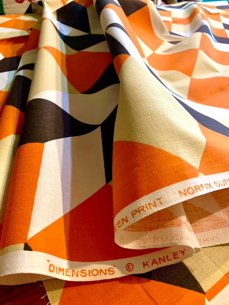 K&ouml;nnte beinhalten: Orangefarbener, schwarz-wei&szlig;er geometrischer Stoff mit dem aufgedruckten Text "DIMENSIONS" und "KANLEY" am Rand. Der Stoff ist gefaltet und der Text ist am Rand der Falte sichtbar.