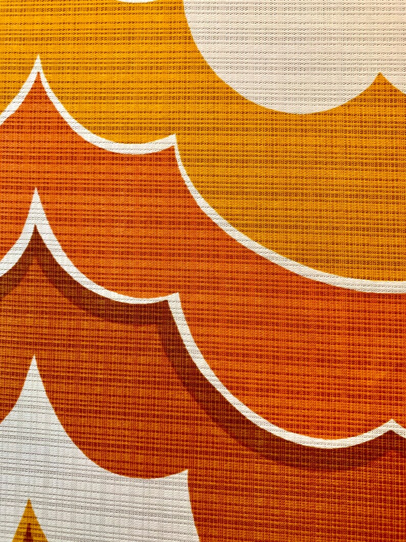 Op de afbeelding: Close-up van een retro stof met een herhalend patroon van gestileerde, gebogen vormen. Het ontwerp heeft tinten oranje, geel en wit, waardoor een getextureerd, gelaagd effect ontstaat. De stof heeft een geweven uiterlijk.