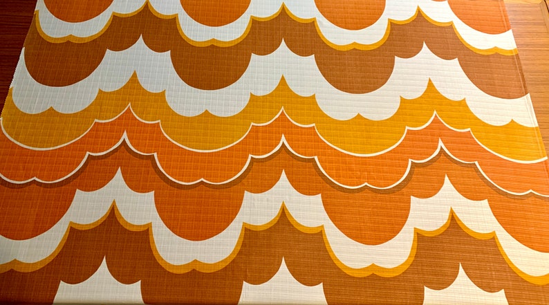 Op de afbeelding: Een stof met een herhalend patroon van gestileerde, wolkvormige vormen in bruin, oranje, geel en wit. Het retro-ontwerp heeft een textuur. De stof is geschikt voor woondecoratie of bekleding.
