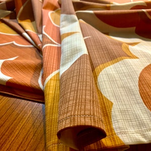 Op de afbeelding: Close-up van een stof met een retro-patroon. Het ontwerp heeft grote, abstracte vormen in tinten bruin, oranje en wit. De stof heeft een getextureerde uitstraling, met zichtbare ribbels en een subtiele glans. De randen zijn netjes genaaid.