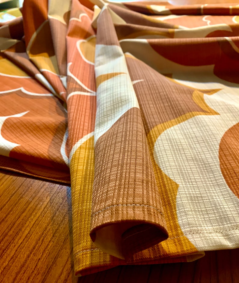 Op de afbeelding: Close-up van een stof met een retro-patroon. Het ontwerp heeft grote, abstracte vormen in tinten bruin, oranje en wit. De stof heeft een getextureerde uitstraling, met zichtbare ribbels en een subtiele glans. De randen zijn netjes genaaid.