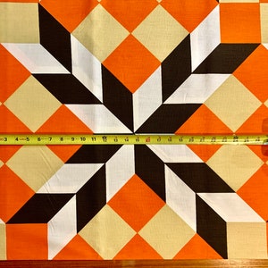 K&ouml;nnte beinhalten: Ein Stoff mit einem geometrischen Muster aus orangefarbenen, wei&szlig;en und braunen Quadraten. Ein Ma&szlig;band liegt auf dem Stoff und zeigt eine Messung von 71 cm.