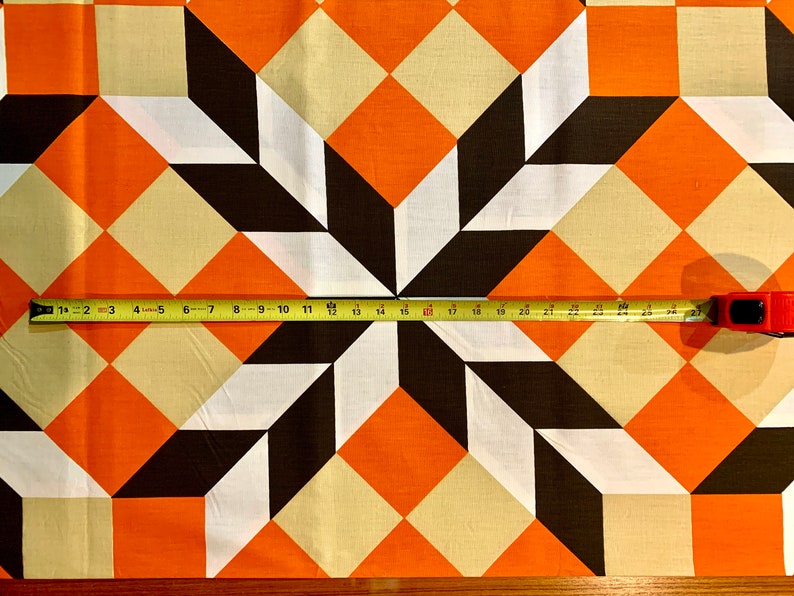 K&ouml;nnte beinhalten: Ein Stoff mit einem geometrischen Muster aus orangefarbenen, wei&szlig;en und braunen Quadraten. Ein Ma&szlig;band liegt auf dem Stoff und zeigt eine Messung von 71 cm.