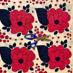 May include: White fabric with a repeating pattern of red roses with dark blue leaves and small red flowers. A pair of blue scissors is placed on the fabric.