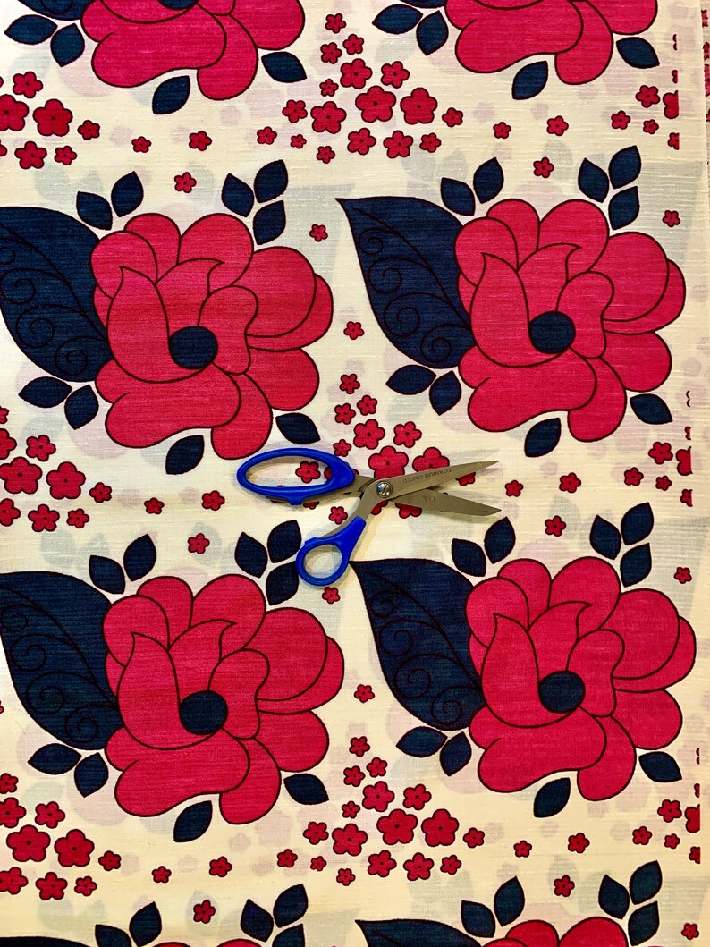 May include: White fabric with a repeating pattern of red roses with dark blue leaves and small red flowers. A pair of blue scissors is placed on the fabric.