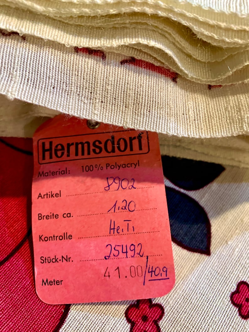 May include: A pink fabric label with black text that reads "Hermsdorf Material: 100% Polyacryl" and includes product information such as "Artikel 8902", "Breite ca. 1.20", "Kontrolle He.T", "St&uuml;ck-Nr. 25492", and "Meter 41.00/409".