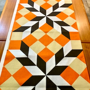 K&ouml;nnte beinhalten: Ein geometrischer Tischl&auml;ufer mit einem sich wiederholenden Muster aus orangefarbenen, schwarzen und wei&szlig;en Rauten und Sternen.