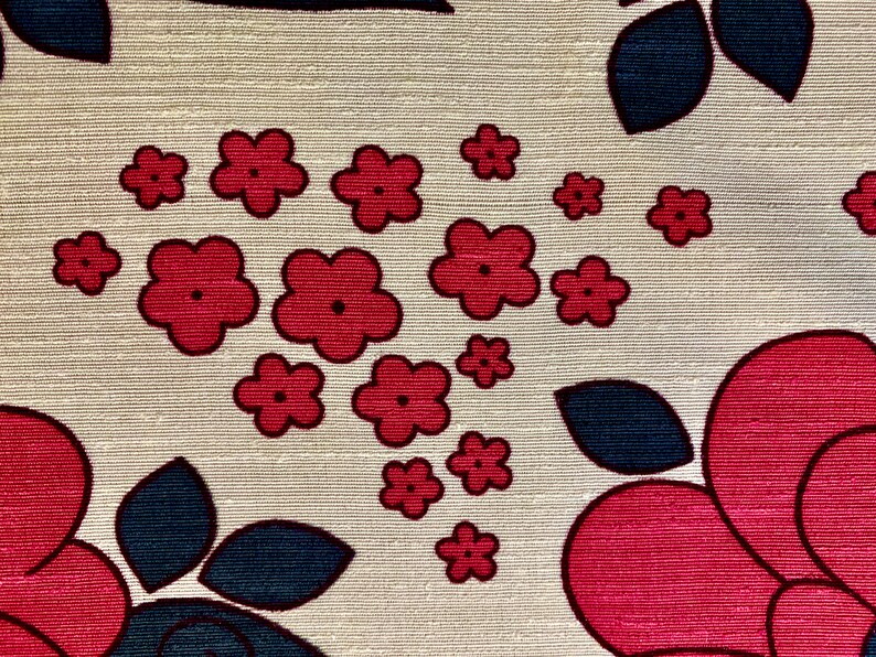 May include: A beige fabric with a repeating pattern of red and blue flowers. The flowers are stylized and have a retro look. The fabric has a textured surface.