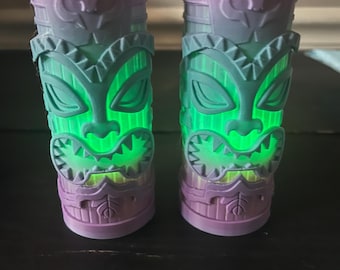 May include: Two purple and teal tiki-style light-up cups. The cups have a tribal design with a skull and geometric shapes. The interior glows green, creating a vibrant effect. The cups are cylindrical and appear to be made of plastic.