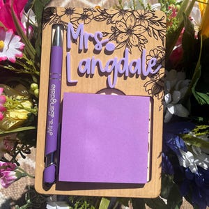 May include: A wooden desk organiser featuring a floral design and the name "Mrs. Langdale" in purple. It includes a purple pen with "Mrs. Langdale" printed on it and a purple sticky note pad.