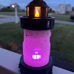 Op de afbeelding: Een decoratief vuurtoren nachtlampje met een zwarte bovenkant en basis, en een roze behuizing. De vuurtoren heeft een oplichtend raam en een warm, flikkerend licht aan de binnenkant van de bovenkant. De behuizing is versierd met zeesterren en schelpen.