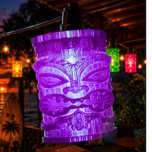 May include: A purple, cylindrical tiki-style light cover featuring a carved face with prominent eyes, a wide nose, and a textured mouth. The translucent material glows from an internal light source, hanging from a black cord as part of a string light set.