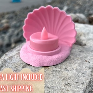 Puede incluir: Un portavelas rosa en forma de concha con una luz de té LED. El soporte tiene una base texturizada y un borde festoneado. La luz de té LED es rosa y tiene forma de llama.