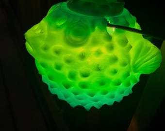 May include: A glowing neon green pufferfish figurine with a textured, spiky surface and a rounded body shape. The object emits a bright, vibrant light, highlighting the detailed scales and fins while casting a soft luminescence in the dark.