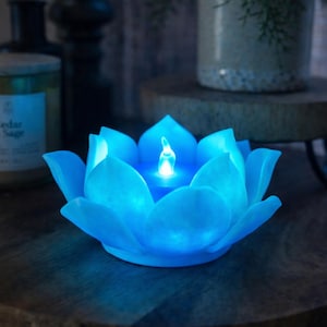 Könnte beinhalten: Eine blaue LED-Kerze in Form einer Lotusblume. Die Kerze hat eine leuchtende Flamme in der Mitte und besteht aus einem durchscheinenden Material. Die Kerze steht auf einer Holzoberfläche.