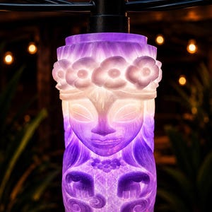 May include: A string of decorative lights featuring a carved tiki-style design. The lights are illuminated with a gradient of purple and white, showcasing a detailed face and floral accents. The string is suspended against a blurred background.
