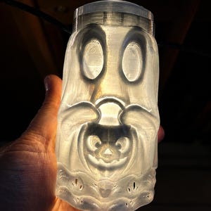 May include: A translucent, 3D-printed cylindrical light cover featuring a relief design of a ghost holding a small jack-o'-lantern. The object is illuminated from within, casting a soft glow that highlights the carved facial features and details.
