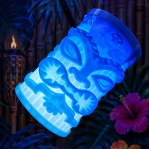 May include: A blue, illuminated tiki-style sculpture with carved facial features and geometric patterns. The sculpture is lit from within, creating a glowing effect. The background features tropical foliage and a torch.