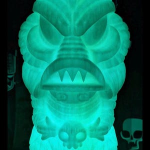 May include: A glowing, translucent green lamp shaped like a creature with a wide, toothy mouth and a pointed head. The figure has textured, layered features and holds a small skull-like object in its hands, emitting a bright, neon-like light.