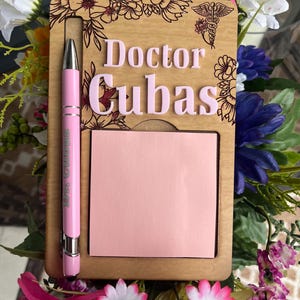 May include: A wooden desk organizer with the name "Doctor Cubas" in pink letters. The organizer includes a pink pen and a pink notepad. Floral designs and a medical symbol are etched into the wood.