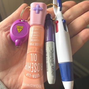 Op de afbeelding: Een verzameling alledaagse voorwerpen, waaronder een paarse sleutelhanger, een roze lipgloss tube met "ROSEHIP OIL" tekst, een paarse Sharpie Mini marker en een witte multi-color pen met blauwe accenten. De lipgloss is 1,2 cm lang.