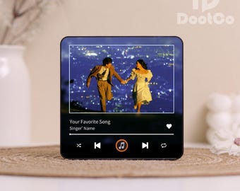 May include: A black rectangular music player displays a romantic image of a couple holding hands. The screen shows a song title and artist name. The player has playback controls and a heart icon. The device is on a woven mat.