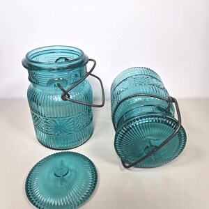 May include: Three turquoise glass jars with lids and metal handles. The jars have a textured, embossed design and are in various positions. One jar is upright, another is on its side, and the lid is separate.