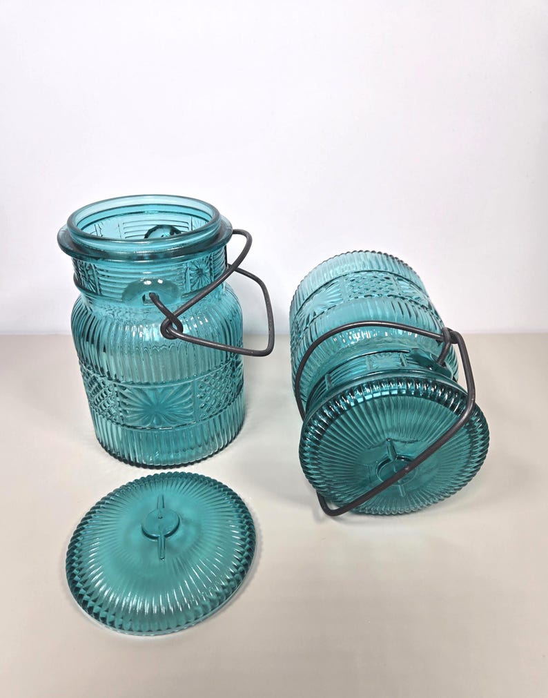 May include: Three turquoise glass jars with lids and metal handles. The jars have a textured, embossed design and are in various positions. One jar is upright, another is on its side, and the lid is separate.