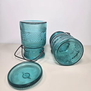 May include: Three-piece teal glass jar set. The set includes two cylindrical jars with textured patterns and metal handles, and a matching lid. The jars are made of teal-colored glass and have a vintage aesthetic.