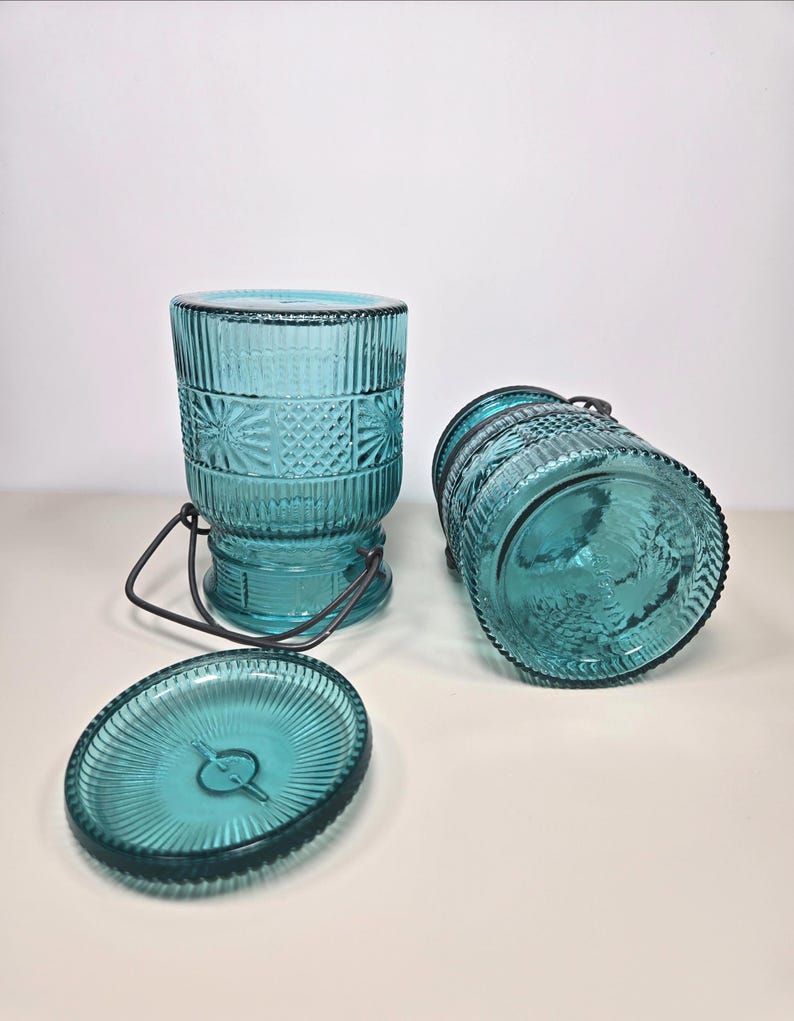 May include: Three-piece teal glass jar set. The set includes two cylindrical jars with textured patterns and metal handles, and a matching lid. The jars are made of teal-colored glass and have a vintage aesthetic.