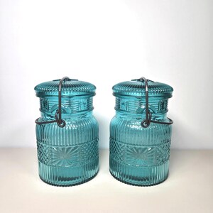 May include: Two teal-colored glass jars with lids and metal handles. The jars have a ribbed design with a decorative pattern around the middle. The lids are also ribbed. The jars are cylindrical and appear to be vintage or antique.
