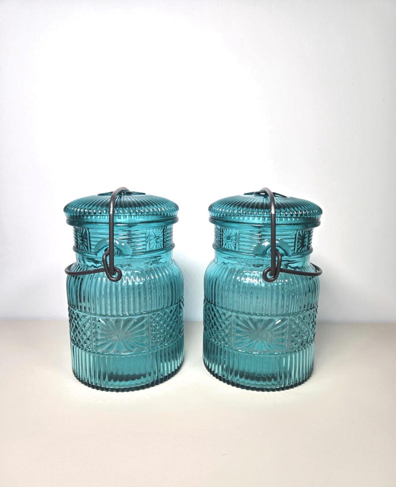 May include: Two teal-colored glass jars with lids and metal handles. The jars have a ribbed design with a decorative pattern around the middle. The lids are also ribbed. The jars are cylindrical and appear to be vintage or antique.