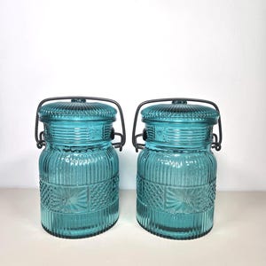May include: Two teal-colored glass jars with ribbed and patterned designs. Each jar has a matching lid and a black metal handle. The jars are likely for storage or decorative purposes.