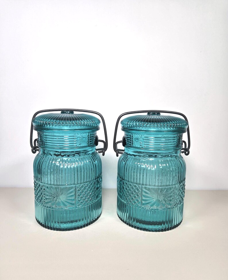 May include: Two teal-colored glass jars with ribbed and patterned designs. Each jar has a matching lid and a black metal handle. The jars are likely for storage or decorative purposes.