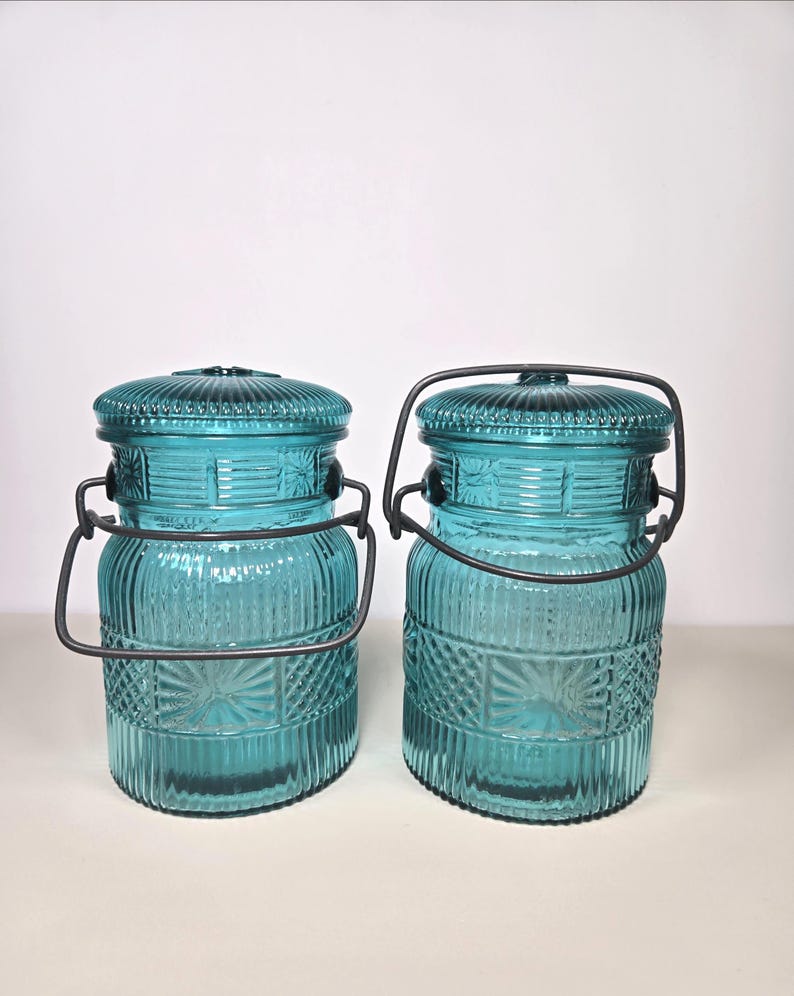 May include: Two matching turquoise glass jars with lids and metal handles. The jars have a ribbed design with decorative starburst patterns. The lids are also ribbed and have a small knob. The metal handles are dark gray.