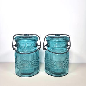 May include: Two matching turquoise glass jars with lids and metal handles. The jars have a textured design with vertical and geometric patterns. The lids are ribbed, and the handles are dark metal. The jars are on a white surface.