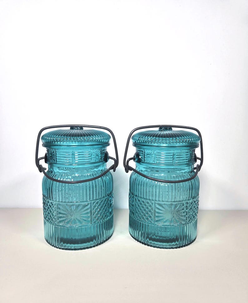 May include: Two matching turquoise glass jars with lids and metal handles. The jars have a textured design with vertical and geometric patterns. The lids are ribbed, and the handles are dark metal. The jars are on a white surface.