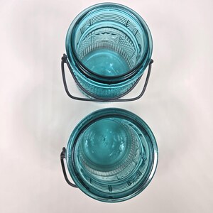 May include: Two turquoise-colored glass jars with decorative patterns and metal handles. The jars are empty and appear to be vintage or antique. The jars are sitting on a white surface.