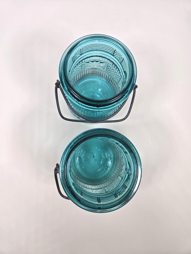 May include: Two turquoise-colored glass jars with decorative patterns and metal handles. The jars are empty and appear to be vintage or antique. The jars are sitting on a white surface.