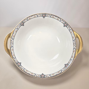 May include: A white serving bowl with gold handles and a decorative floral border in blue, pink, and gold. The bowl has a wide, shallow design, suitable for serving dishes.