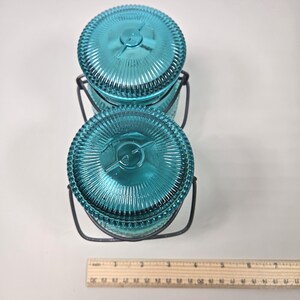 May include: Two teal glass jars with ribbed lids and metal handles. The jars are stacked, and a ruler is placed next to them. The jars are approximately 3 inches in diameter.