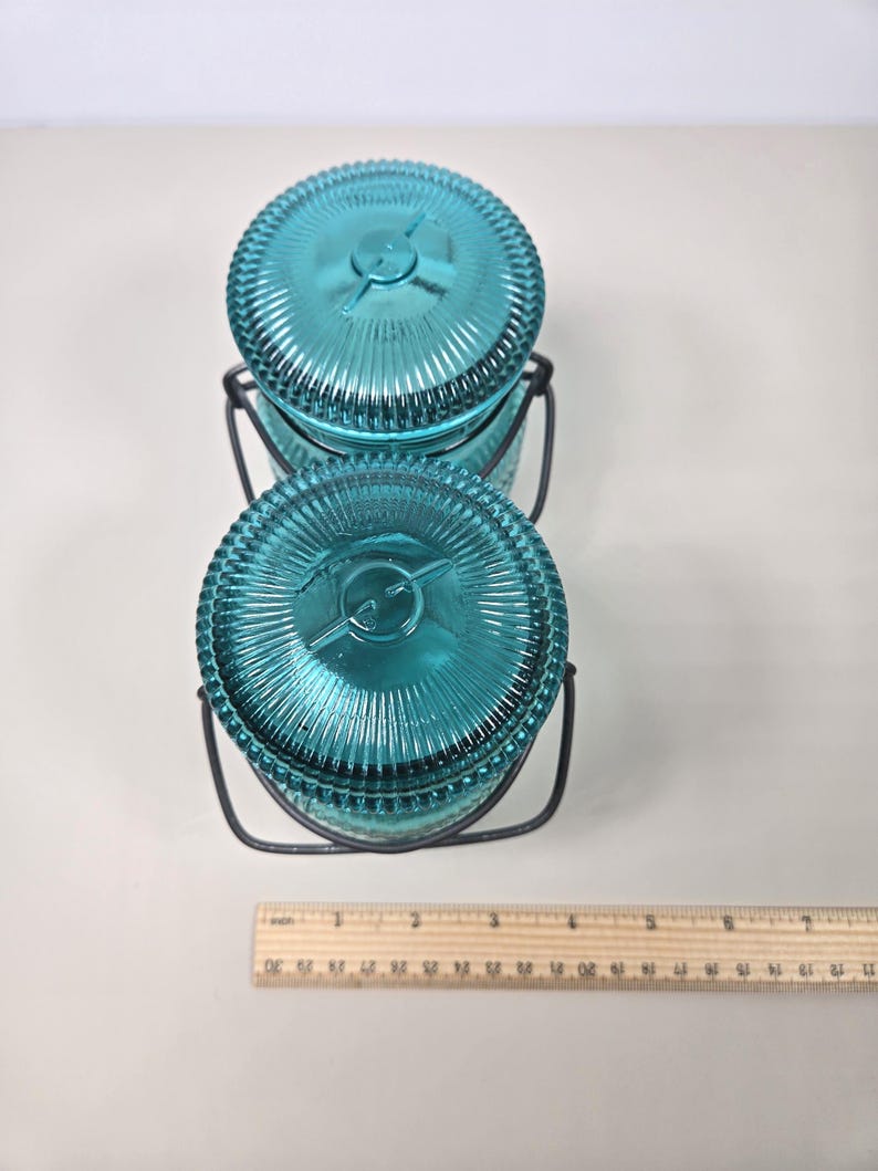 May include: Two teal glass jars with ribbed lids and metal handles. The jars are stacked, and a ruler is placed next to them. The jars are approximately 3 inches in diameter.