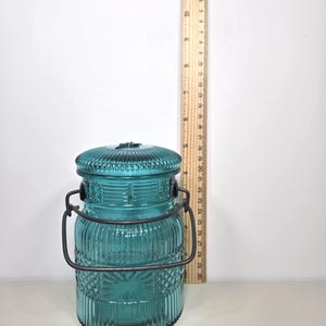 May include: A teal glass jar with a lid and a metal handle. The jar has a ribbed and patterned design. A wooden ruler is next to the jar, measuring approximately 5 inches in height.