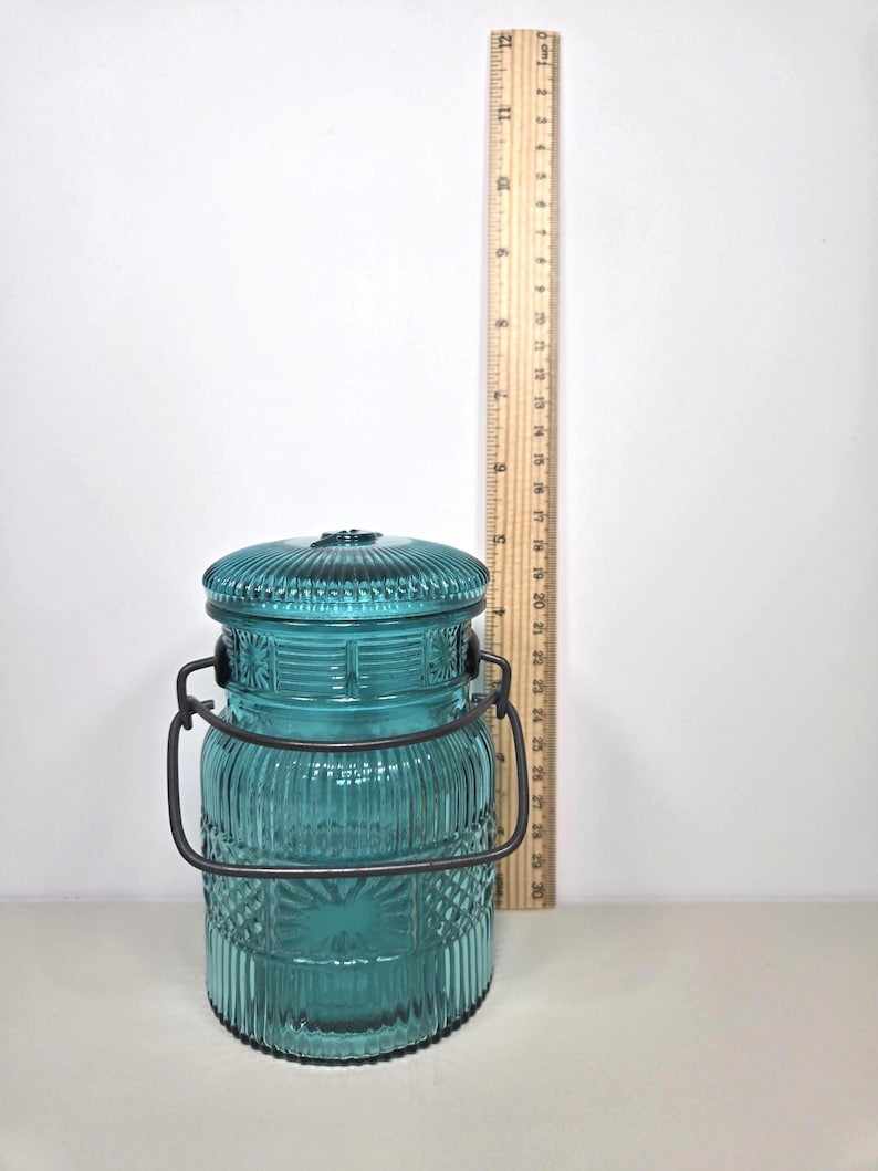 May include: A teal glass jar with a lid and a metal handle. The jar has a ribbed and patterned design. A wooden ruler is next to the jar, measuring approximately 5 inches in height.