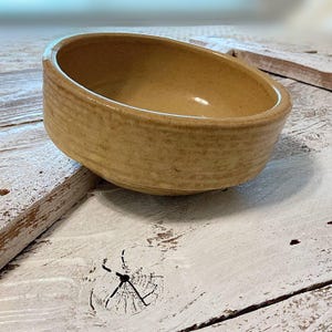 May include: A small, round, yellow ceramic bowl with a textured surface. The bowl is sitting on a white wooden surface.