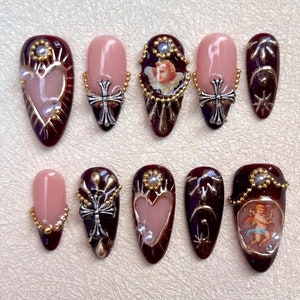 Könnte beinhalten: Satz von zehn falschen Nägeln mit verschiedenen Designs. Die Designs beinhalten ein Herz, ein Kreuz und einen Engel. Die Nägel sind burgunderrot mit goldenen Akzenten.