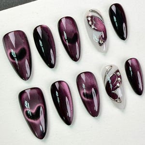 May include: A set of ten almond-shaped press-on nails featuring a deep purple cat-eye magnetic polish finish. Two of the nails have a white base with a purple butterfly wing design accented by small silver rhinestones and metallic silver lines.