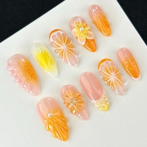 Könnte beinhalten: Eine Sammlung künstlicher Fingernägel mit verschiedenen Designs. Einige Nägel zeigen Orangenscheiben, während andere florale und abstrakte Muster in Orange-, Gelb- und Rosatönen aufweisen. Mehrere Nägel sind mit kleinen, glitzernden Verzierungen versehen.