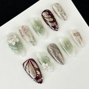 Puede incluir: Una colección de diez uñas artificiales con diversos diseños. Las uñas presentan colores borgoña, plateado y verde, con patrones florales y abstractos. Algunas uñas tienen acentos florales en 3D, mientras que otras muestran detalles de purpurina y oro. Cada uña mide unos 2 cm.
