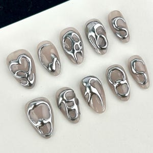 Könnte beinhalten: Ein Set aus zehn mandelförmigen Kunstnägeln. Jeder Nagel ist in einem Nudeton gehalten und mit einem silbernen, abstrakten Design versehen. Die silbernen Designs variieren, darunter Wirbel und organische Formen, für einen modernen Look.